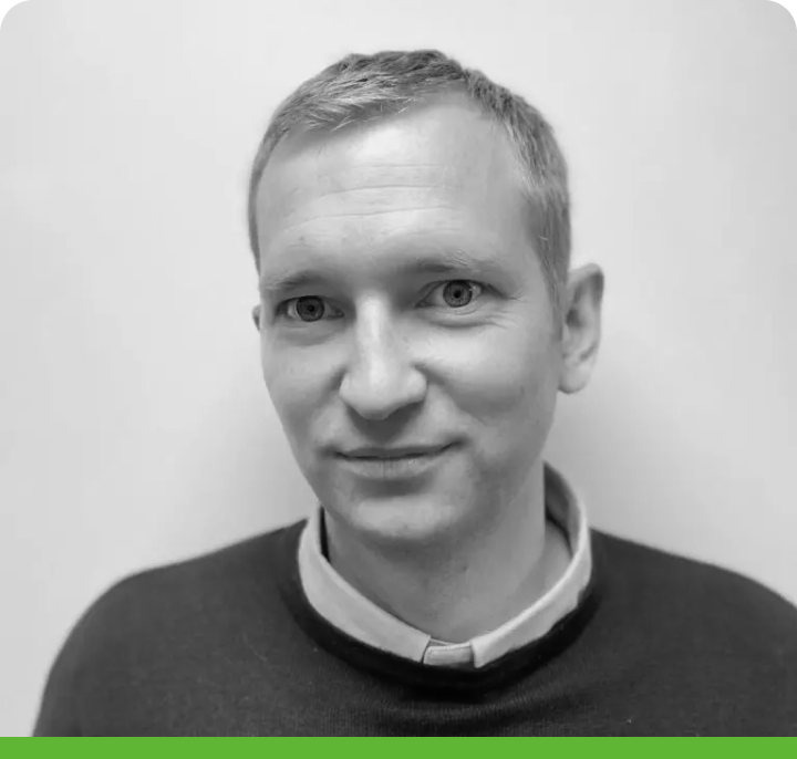 Black and white photo of a person with short hair in a collared shirt under a sweater, against a plain background; green border at the bottom.