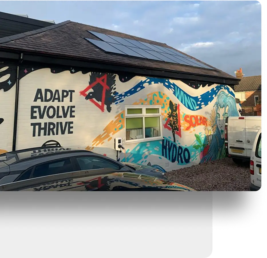 Mural on building wall shows waves, solar panels, wind turbines, and words: ADAPT, EVOLVE, THRIVE, SOLAR, HYDRO. Cars parked nearby; solar panels on roof.