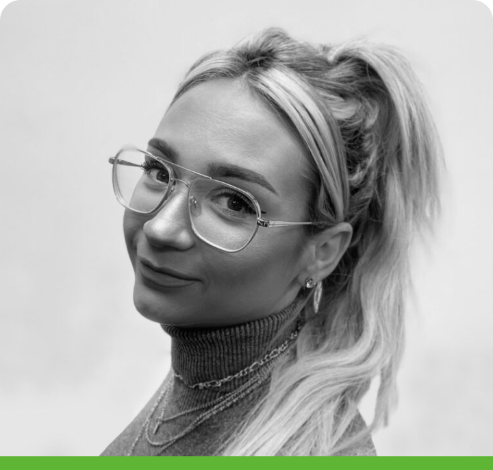 Black and white portrait of a smiling person with long blonde hair in a high ponytail, wearing glasses and a turtleneck, looking over their shoulder against a plain background.