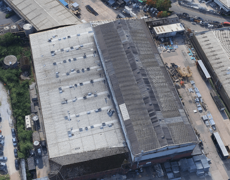 Aerial view of an industrial warehouse with a corrugated metal roof, surrounded by construction materials, storage containers, greenery, roads, and other buildings.