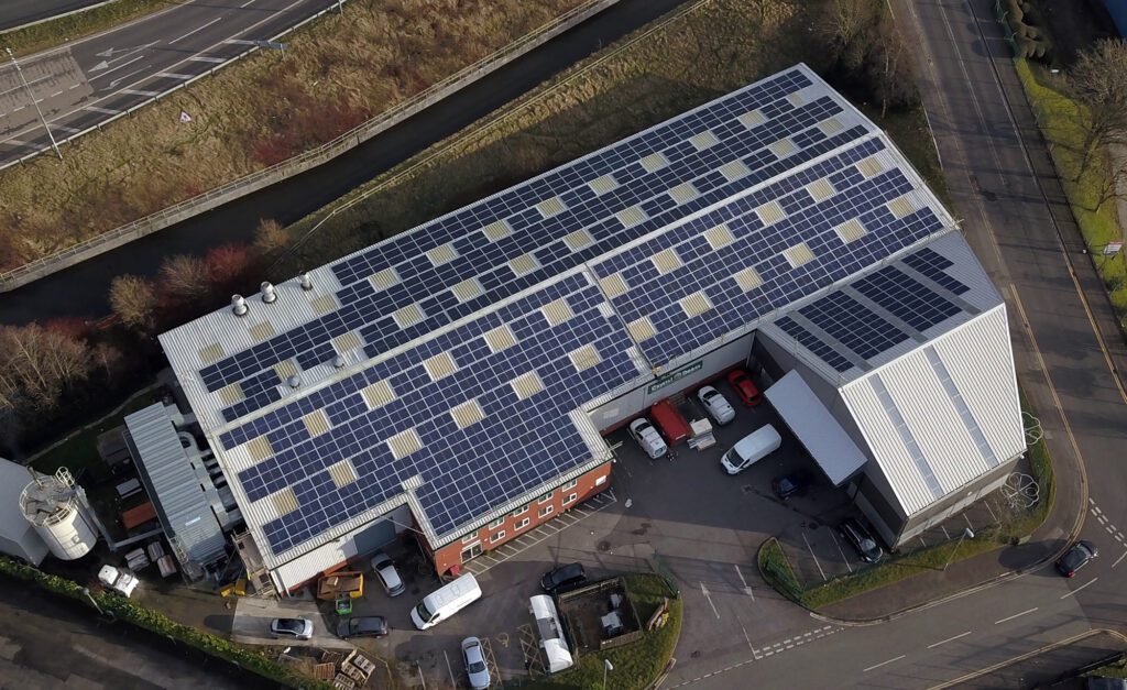 Aerial view of an industrial building with solar panels covering the entire rooftop, surrounded by parked vehicles.