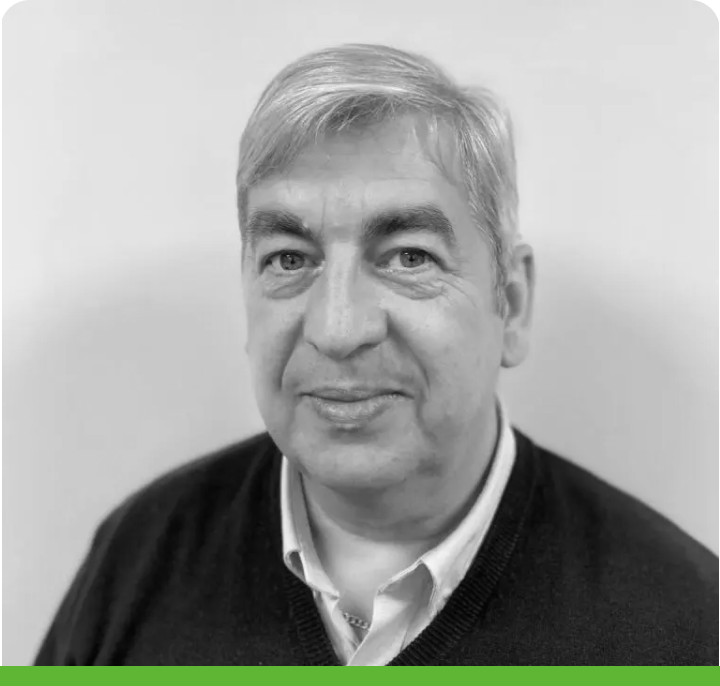 Grayscale portrait of an older man with short gray hair, wearing a dark sweater over a collared shirt. He looks directly at the camera against a plain background with a green border at the bottom.