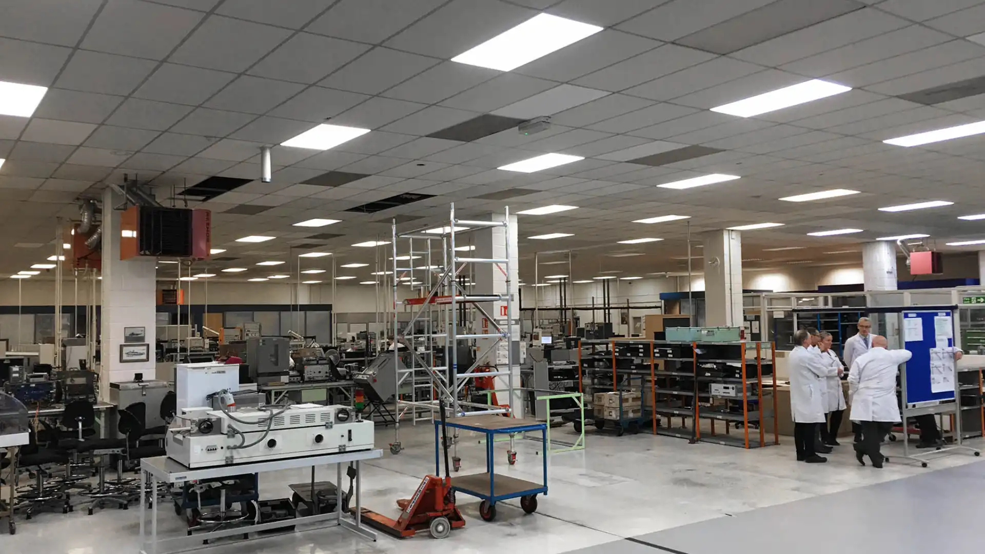 Expansive, well-lit lab by AT Engineering Solutions with machinery and a central metal scaffolding tower. Two people in white lab coats converse on the right.