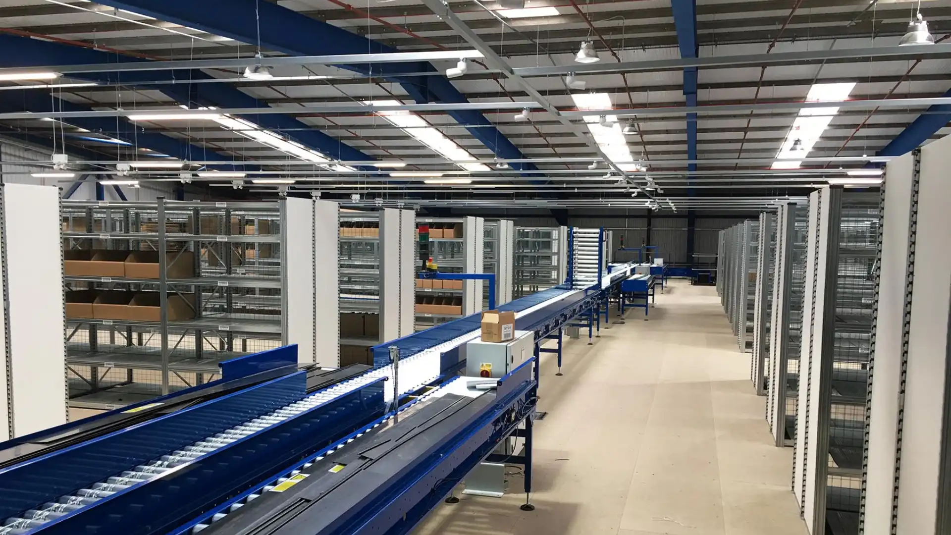 Interior of a spacious warehouse featuring blue conveyor belts and tall metal shelves. Boxes are on the conveyors, and the area is brightly lit with overhead lights. Exposed beams and ductwork adorn the ceiling.
