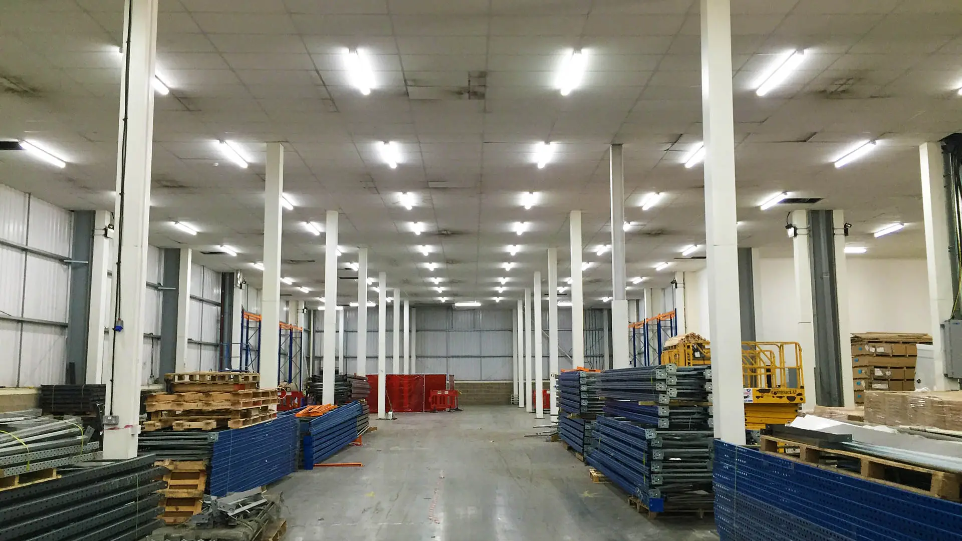 Spacious Unilin warehouse interior with high ceilings, blue and orange metal shelves, wooden pallets, and a clear central path, showcasing a clean and organized space.