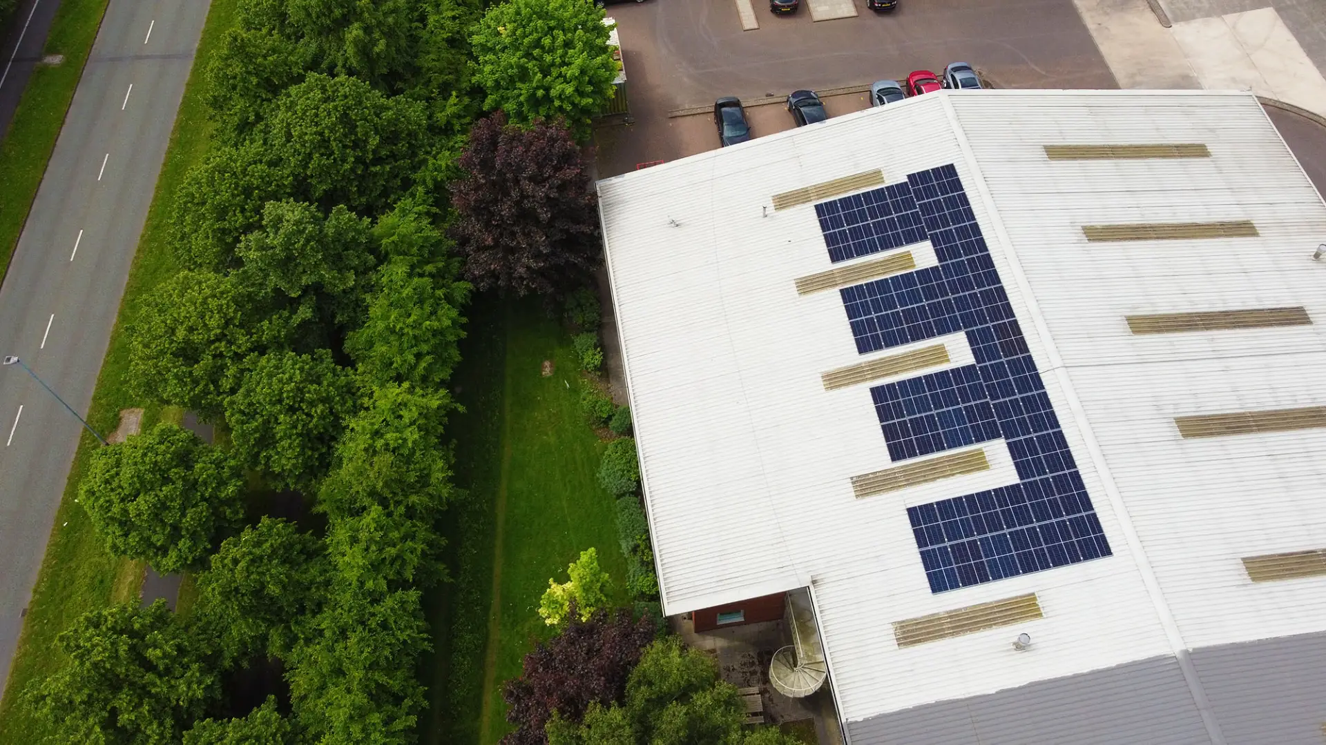 Building with rooftop solar panels, surrounded by cars, trees, and green landscape.