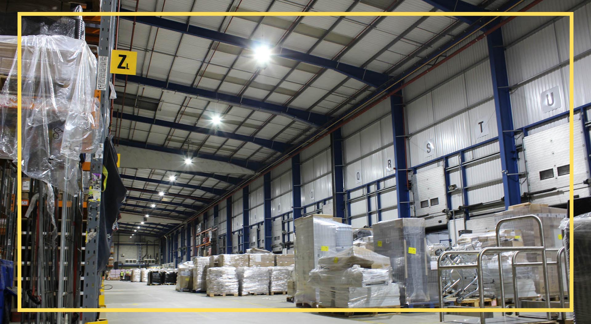 Interior of a spacious warehouse with high ceilings, bright overhead lighting, and metal beams. The area features multiple labeled loading dock doors and stacks of pallets wrapped in plastic containing various goods. Shelves on the left side hold items covered in plastic, while a sign with 'ZL' hangs from one of the shelves, emphasizing organized storage and logistics.