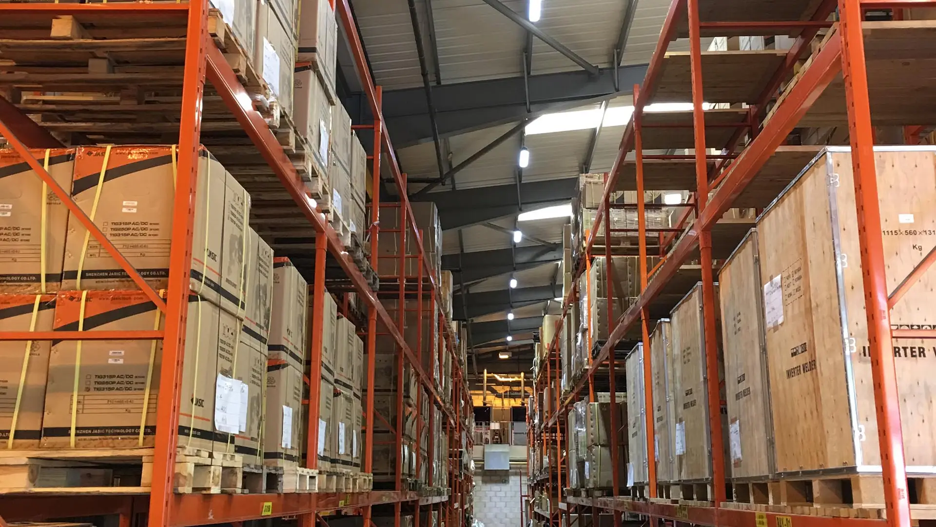 Warehouse interior with tall orange metal shelves filled with cardboard boxes and wooden crates under bright lights.