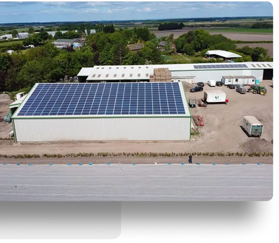 Aerial view of a large white industrial building with solar panels on the roof. Surrounding the building are trees, fields, and additional structures, with a road in the foreground. Equipment and vehicles are visible next to the building.