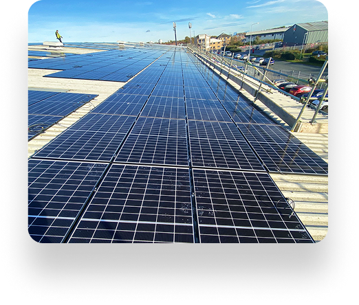 Wide view of rooftop solar panels under a clear blue sky, aligned by AK Sheet Metal Fabrications. Panels reflect sunlight. Buildings, cars, and greenery in background. A person stands on the roof afar.
