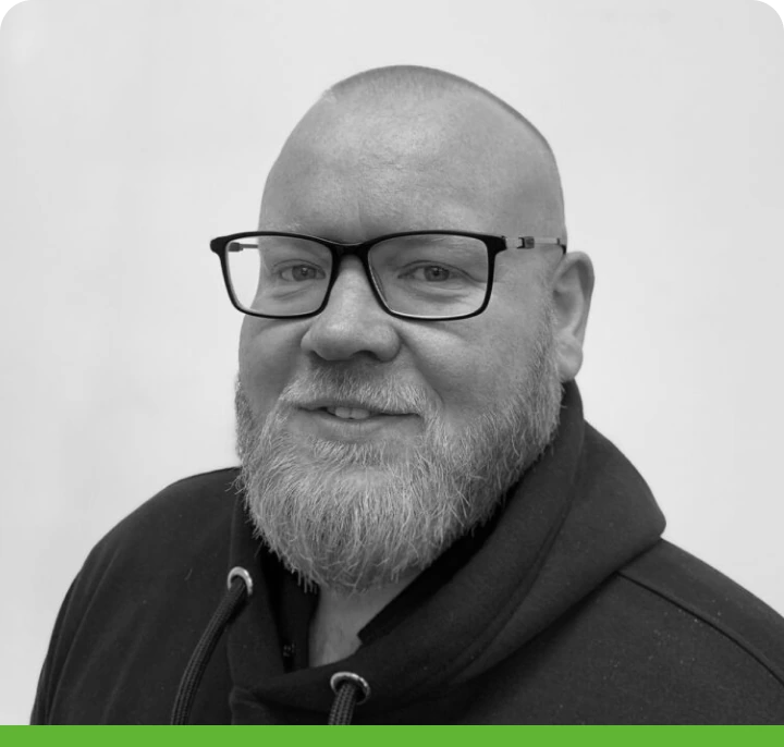 A man with a beard and glasses smiles slightly, wearing a hoodie. The image is black and white with a rounded border and green footer.