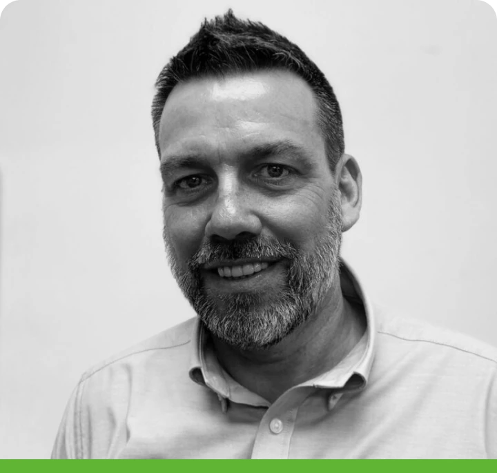 Black and white photo of a smiling man with short dark hair and a beard, wearing a light button-up shirt. Green bar at the bottom of the image.