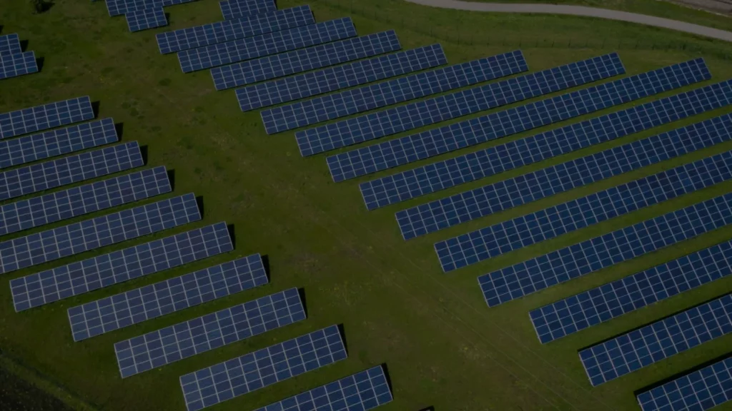 Aerial view of a solar farm with rows of blue panels on a grassy field. A paved path runs along the top edge.