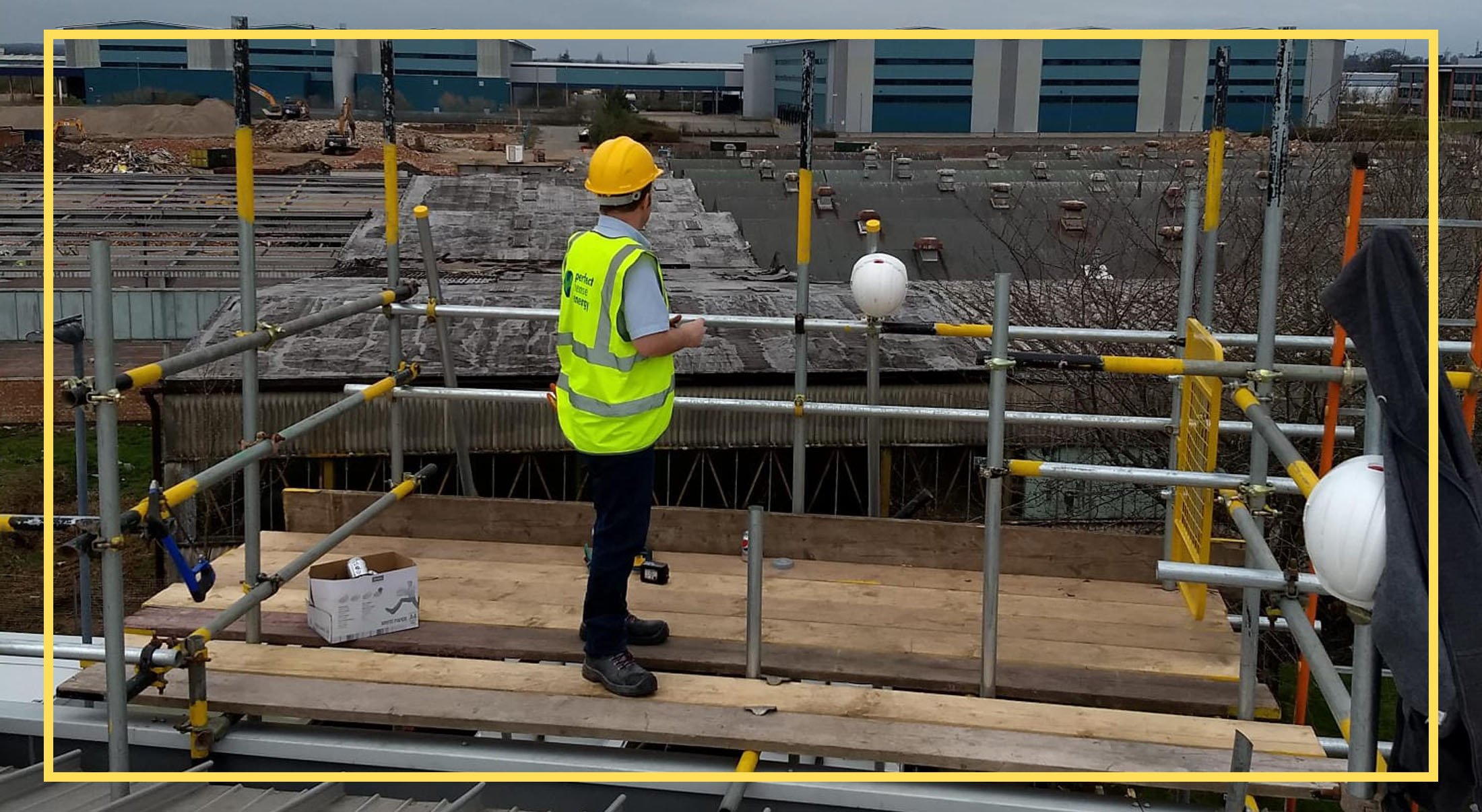 Worker in safety gear standing on scaffold at a construction site, buildings in background.