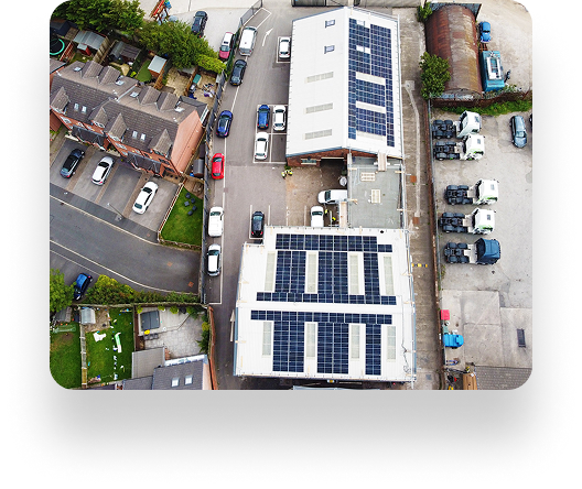 Aerial view of a commercial area with two large buildings having solar panels on rooftops. Vehicles and parked trucks surround the buildings, near residential houses.