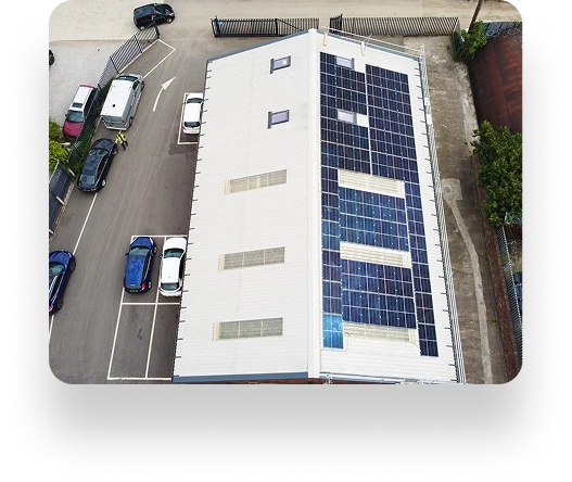 Aerial view of a rectangular industrial building with solar panels on the roof, surrounded by parked cars, paved ground, and a fenced area in the background.
