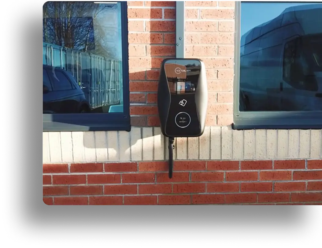 Wall-mounted electric vehicle charging station with a cable, installed on a red brick wall between two windows. Reflections of trees and a vehicle are visible in the windows.