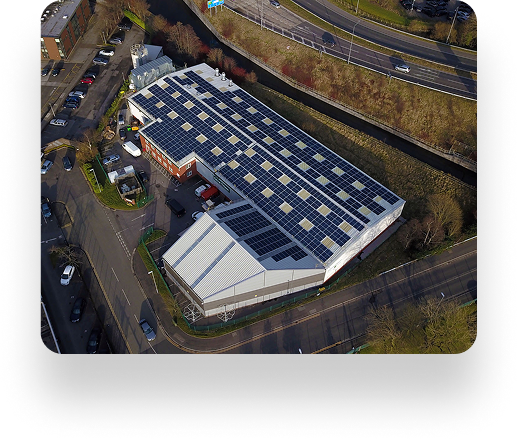 Aerial view of a large industrial building with solar panels and Crystal Doors, surrounded by roads, parked cars, lush greenery, and a scenic canal.