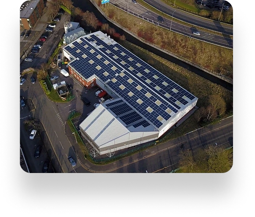 Aerial view of a large industrial building with solar panels and Crystal Doors, surrounded by roads, parked cars, lush greenery, and a scenic canal.