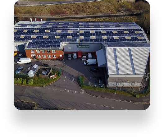 Aerial view of an industrial building with solar panels on the roof, surrounded by a parking lot with cars. A road curves nearby, and another building is adjacent.