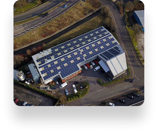 Aerial view of a large industrial building with solar panels on the roof. Crystal doors are surrounded by parked vehicles, roads, and lush greenery.
