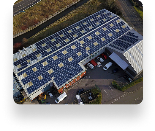Aerial view of a large industrial building with solar panels on the roof. Trucks and vans are parked outside, and roads weave through lush greenery nearby.