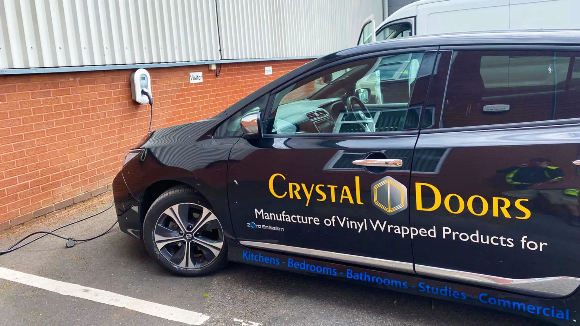 A sleek black car with the Crystal Doors logo charges at an EV station by a brick wall. The side displays Manufacture of Vinyl Wrapped Products, symbolizing innovation and sophistication.