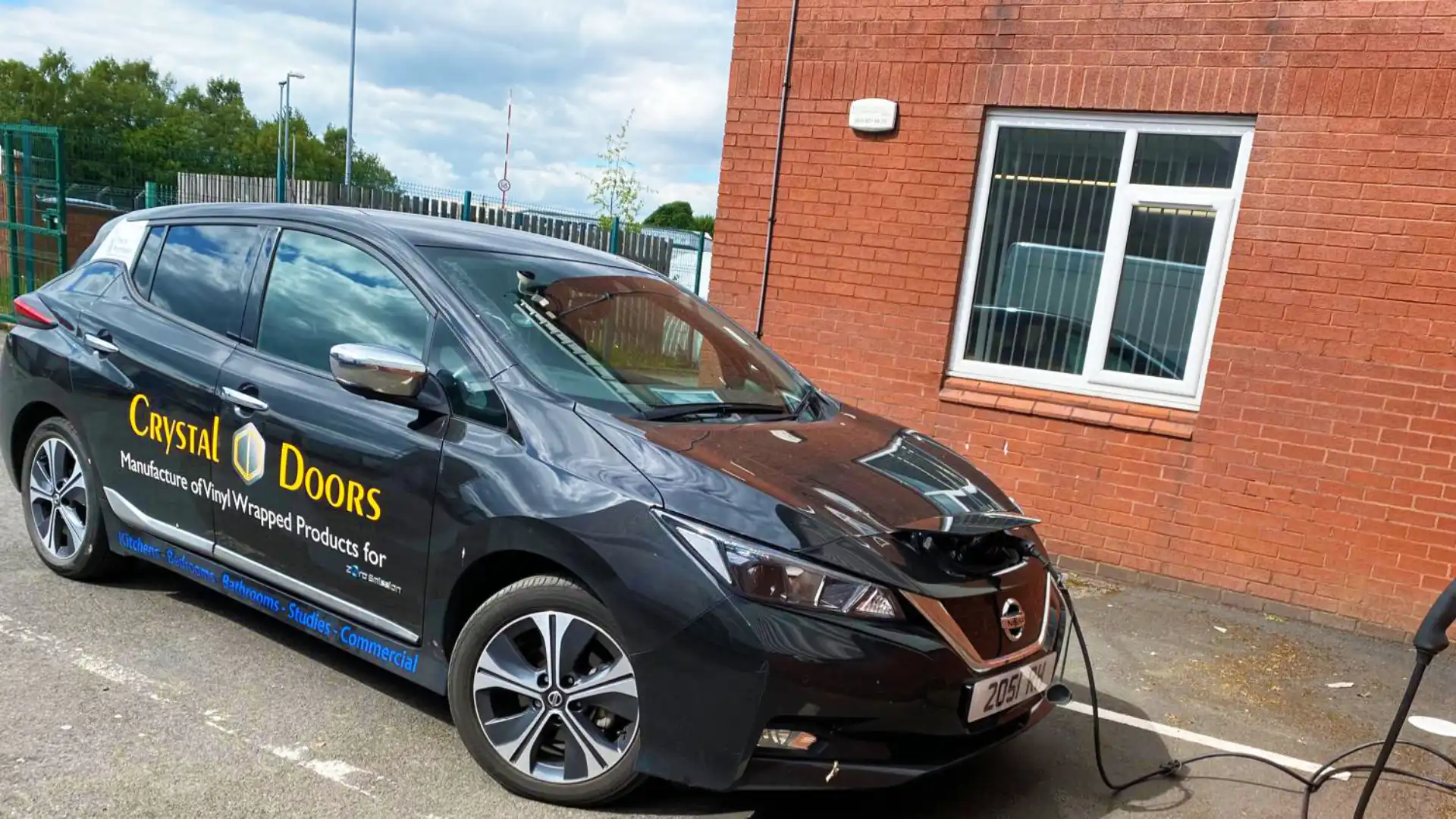 Electric car from Crystal Doors charging outside a brick building, displaying company branding. Cable connects to front. Parking area with windows and railings in the background.