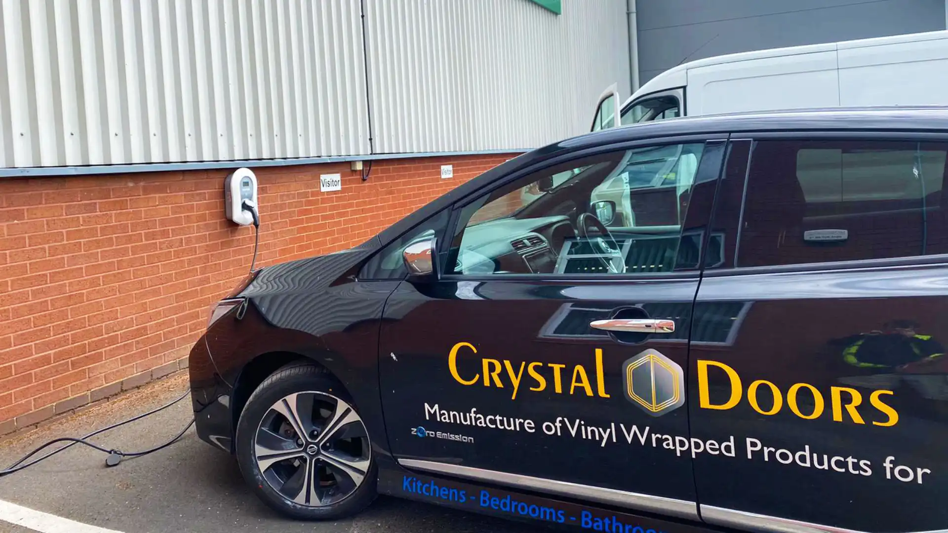 A sleek black electric car with the Crystal Doors logo charges at a wall-mounted station, highlighting expertise in vinyl-wrapped home products. A white van is parked nearby.