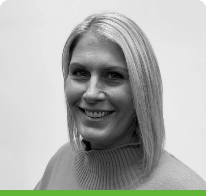 Black and white portrait of a smiling woman with shoulder-length blonde hair. She is wearing a ribbed sweater. The photo features rounded corners and a green bottom border.