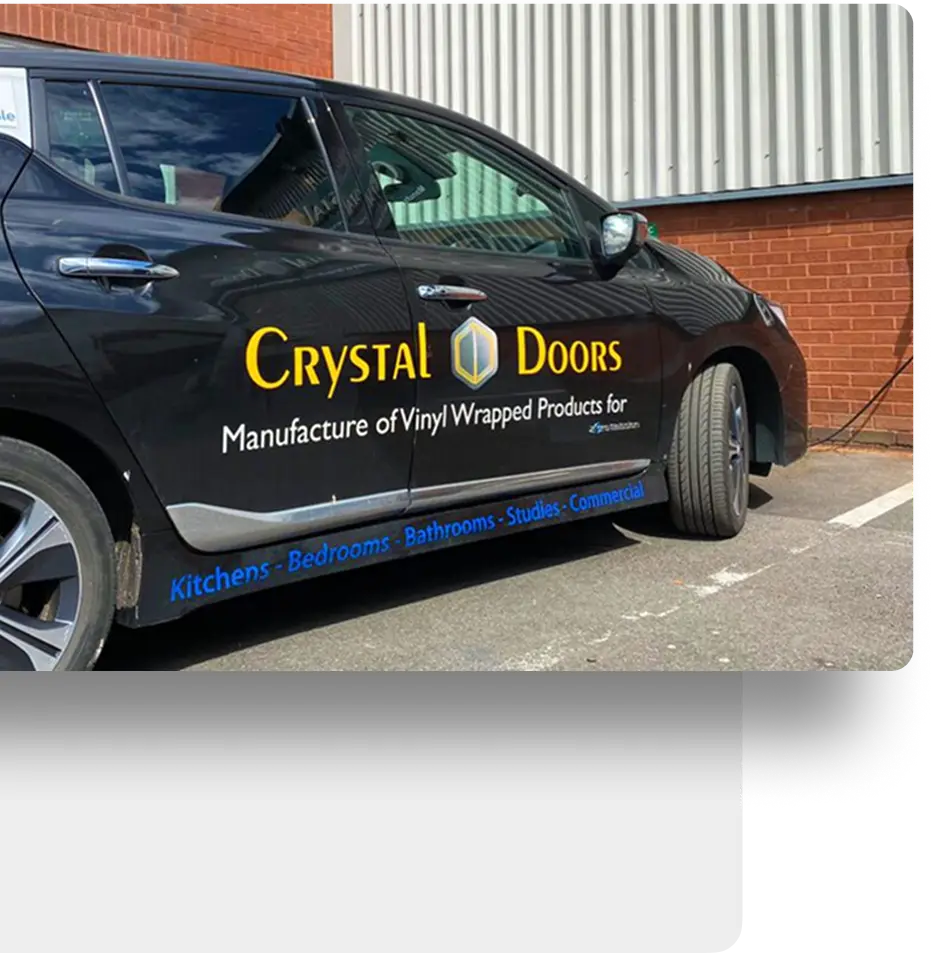 A dark-colored car is parked outside a brick building. The cars door features an advertisement for Crystal Doors, a company specializing in vinyl-wrapped products for kitchens, bedrooms, bathrooms, and commercial spaces.