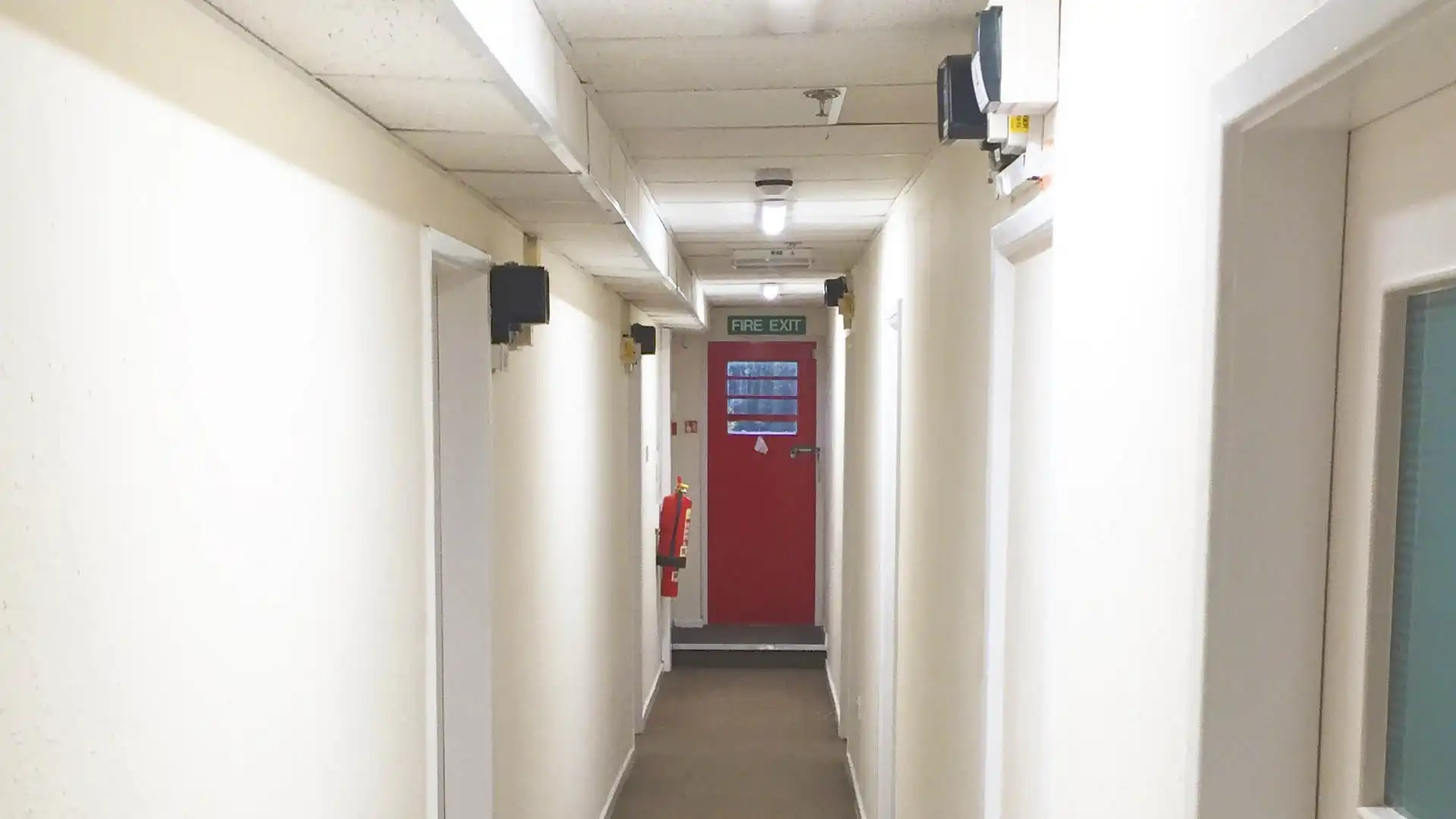 Hallway with white walls, beige carpet, light fixtures, fire extinguisher, and a red fire exit door.