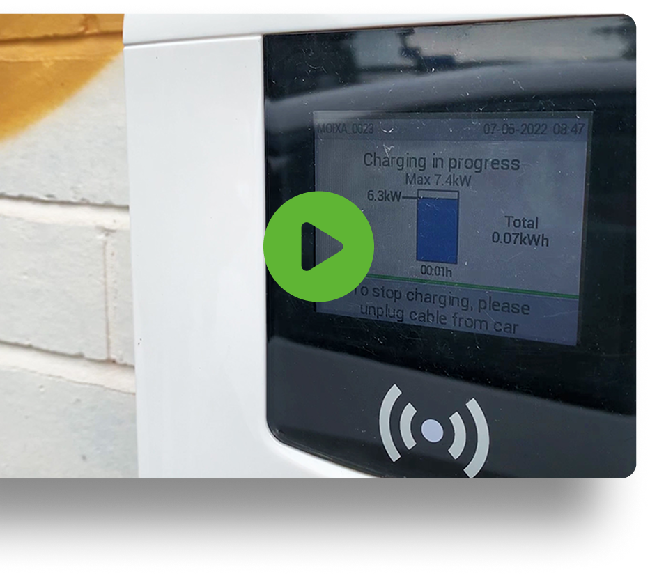 Close-up of a charging station display showing Charging in progress with a maximum of 7.4 kW. The total energy used is 0.7 kWh. A green play button is overlaid on the image, and the background shows a white brick wall with a yellow stripe.
