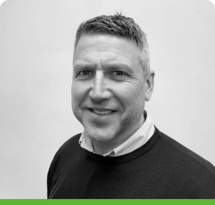 Black and white portrait of a smiling man with short hair, in a dark sweater over a light collared shirt. Plain white background with a green bar at the bottom.