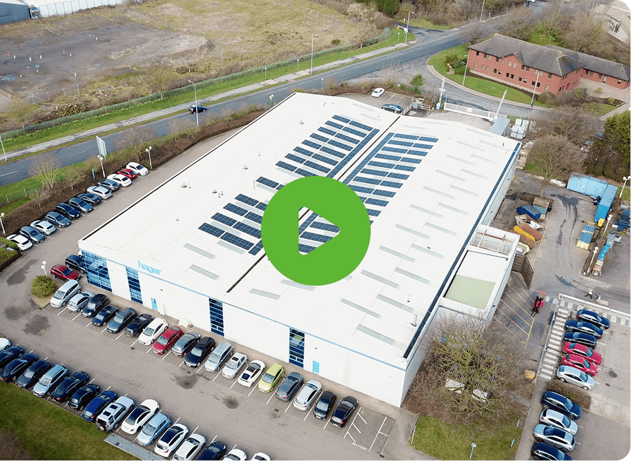 Aerial view of Hager UKs large industrial building with solar panels on the roof. Cars fill the parking lot. A green play button icon suggests a video presence.