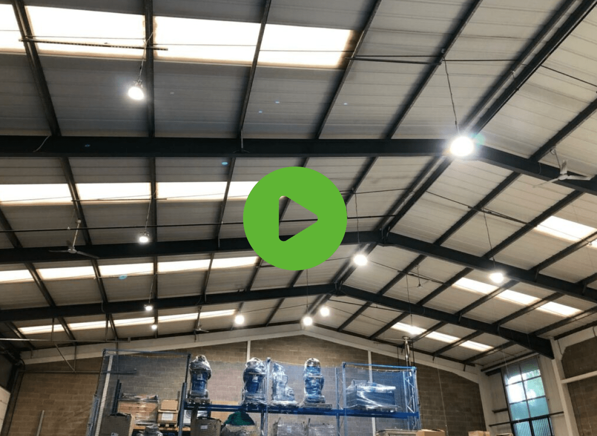 A spacious warehouse with a high metal ceiling and bright lights. Shelves filled with wrapped goods line the walls. A large green play button is in the center.
