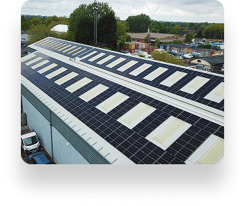 Aerial view of a large industrial building with solar panels on its roof, surrounded by trees and other buildings, highlighting green energy in an industrial area.