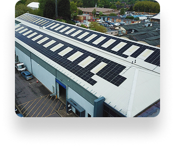 Aerial view of a large industrial building with blue accents and many solar panels on the roof, surrounded by trees and a parking area under an overcast sky.