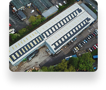 Aerial view of a large industrial building with a white roof and solar panels, surrounded by parked cars, trees, adjacent structures, and roads.