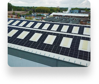 A large industrial building with a solar panel-covered roof. Nearby buildings and smoke in the distance are visible under a partly cloudy sky.