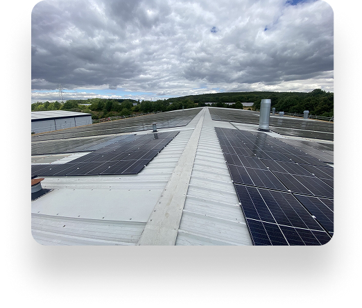 A flat rooftop with solar panels extends towards the horizon under a partly cloudy sky, surrounded by trees and industrial buildings in the distance.