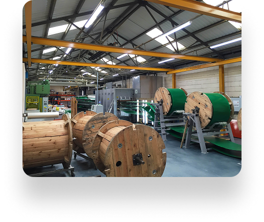 Factory interior with wooden cable reels on the floor, green Kadant machinery in the background, bright overhead lights, and a metal roof structure.