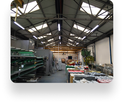 The Kadant-equipped warehouse interior showcases industrial machines, materials stacks, and efficient lighting. Green conveyor belts merge with silver machinery on the left; neatly packaged goods are on the right. The high ceiling features a sturdy metal framework and skylights.