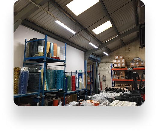 The Kadant warehouse has metal shelves with colorful material rolls. Boxes are stacked on a back shelf, and wrapped items are on the floor. Bright overhead lights illuminate the organized space.