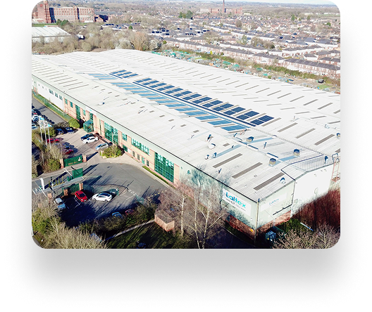 Aerial view of Laltex Groups large warehouse with a white roof and skylights, surrounded by parked cars and sparse trees. Residential areas are visible in the background.