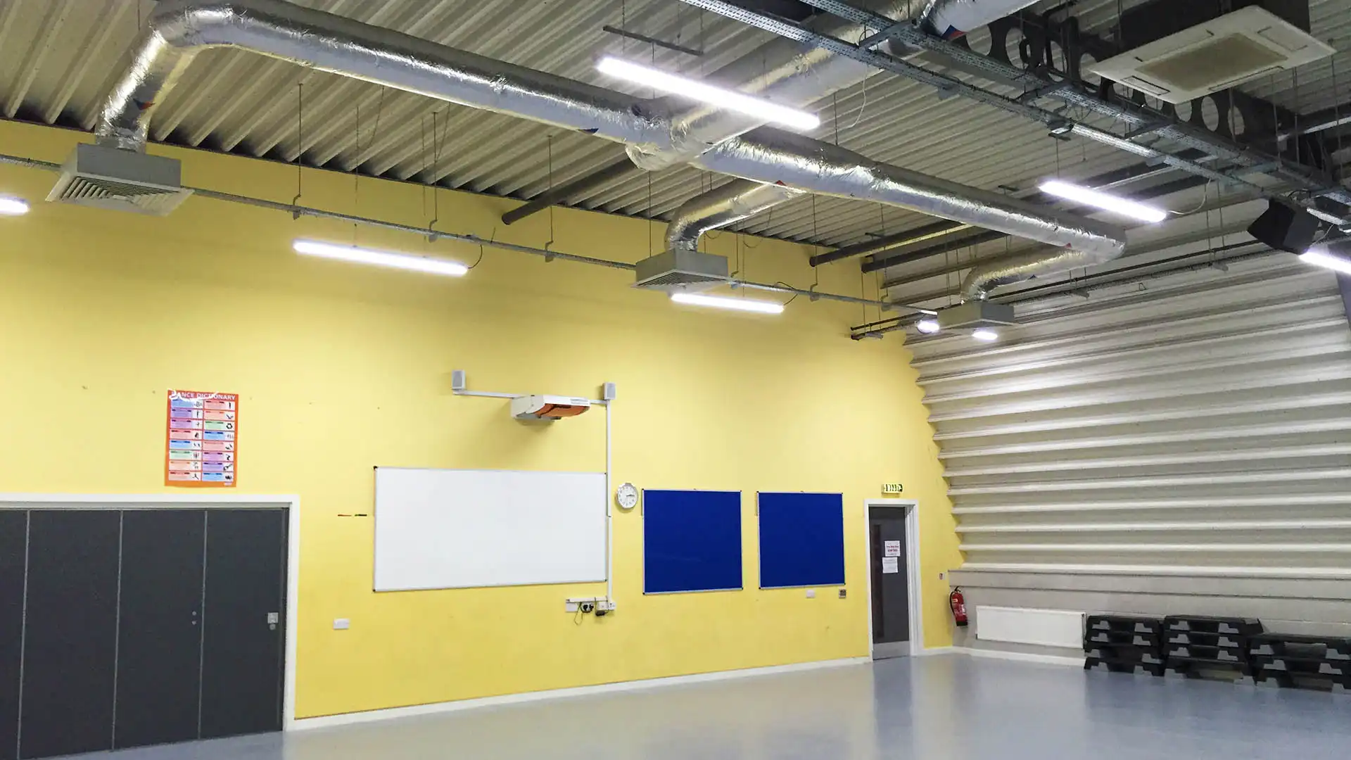 Yellow-walled room with a whiteboard, bulletin boards, chairs, and industrial ceiling.