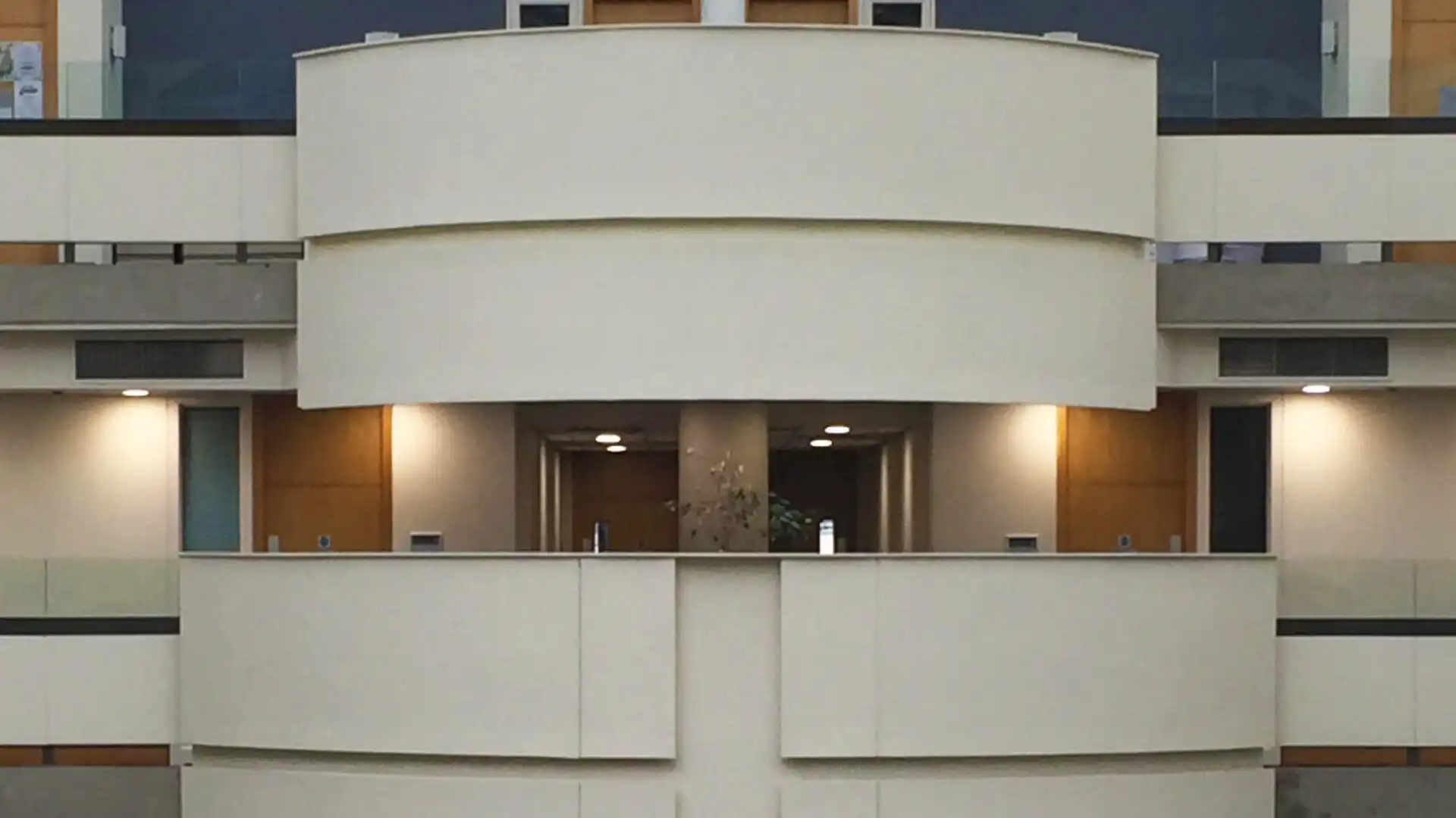 Symmetrical modern architecture resembles Manchester Metropolitan University with beige curved balconies. Three levels have recessed lighting and glass railings, framed by light wood panels on the sides.