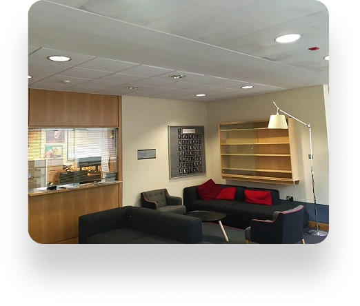 A sleek waiting room features black sofas with red pillows, a round black table, and empty wooden shelves. A floor lamp stands in the corner, with small portraits on the wall. The windowed reception desk is well-lit on the left side.