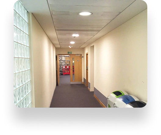 A corridor with beige walls and dark carpet leads to an exit. Recycling bins on the right. Ceiling lights brighten the space, with glass bricks on the left wall.
