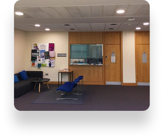 A waiting room at Manchester Metropolitan University with a black sofa, two blue chairs, and a small black table. A bulletin board with posters is on the wall. On the right are two wooden doors, and in the center is a windowed reception area with blinds.